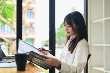 Asyalı kadın finansör kağıt döküman okuyor, pazarlama verilerini parlak ofiste analiz ediyor..