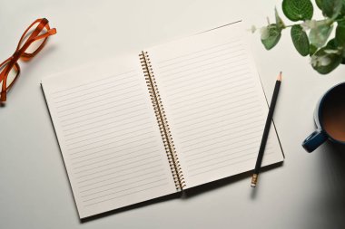 Üst görünüm, düz boş defter, beyaz ofis masasında kalem, bardak ve kahve fincanı..