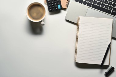 Boş not defteri, dizüstü bilgisayar, hesap makinesi ve kahve fincanı beyaz ofis masasında. Metnin için alanı olan en üstteki görünüm.