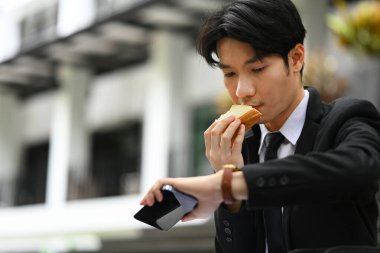 Asyalı erkek ofis çalışanı ofis binasının dışında yemek yiyor, sandviç yiyor ve bilek saatini kontrol ediyor..