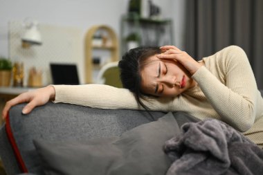 Üzgün genç bir kadın kanepeye uzanıyor ve kafasına dokunuyor. Kronik baş ağrısı çekiyor..