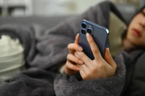 ソファーに横たわり インターネットで医療情報を検索するためにスマートフォンを使用して毛布の病気の女性 — ストック写真