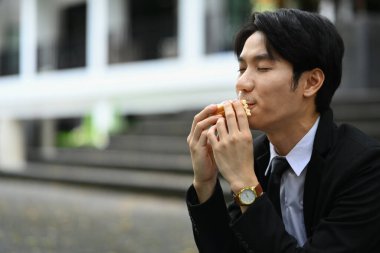 Ofis binasının önünde oturmuş sandviç yiyen mutlu bir erkek ofis çalışanı. Fast food ve şehir hayatı konsepti.