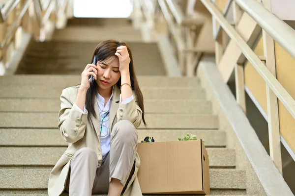 Sad female worker talking on mobile phone while sitting on stairs of building with belongings after being fired from her job.