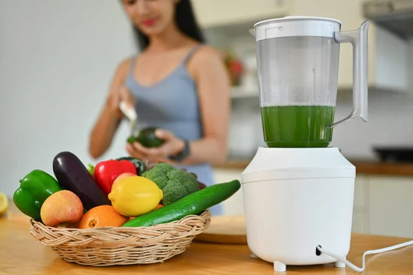 Blender and fresh vegetables on kitchen countertop with young woman cooking in background. Dieting, healthy food concept.