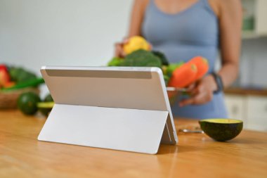 Dijital tablet ve ahşap mutfak tezgahında yarım avokado ve arka planda yemek pişiren genç bir kadın..