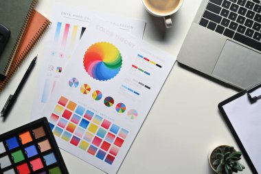 Yaratıcı tasarımcı, laptop, renk örnekleri ve beyaz masadaki kırtasiye malzemeleriyle çalışma alanında grafiksel çizim.