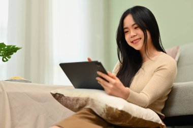 Oturma odasında dijital tablet kullanan sevimli, gülümseyen genç bir kadın. Teknoloji, insanlar ve yaşam tarzı kavramı.