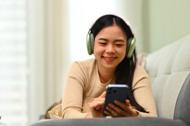 Kulaklıklı neşeli genç bir kadın akıllı telefondan sosyal medyada arkadaşlarıyla sohbet ediyor..