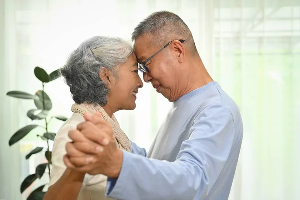 Happy elderly couple spouses dancing in living room while spending time together at home.