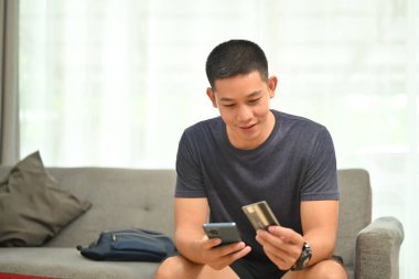 Yakışıklı Asyalı adam kredi kartı tutuyor ve internet üzerinden alışveriş yapmak için cep telefonu kullanıyor..