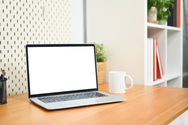 Boş ekranlı dizüstü bilgisayar, kahve fincanı, kalem tutacağı ve oturma odasındaki ahşap masada saksı bitkisi..