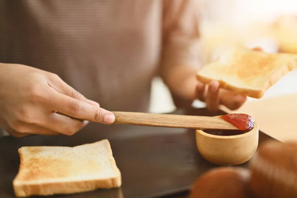 Cropped shot of woman spreading strawberry jam on slice of bread while preparing breakfast in kitchen.