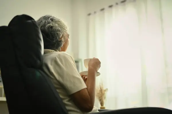 Calm and peaceful elderly woman with cup of coffee sitting on armchair and looking away.