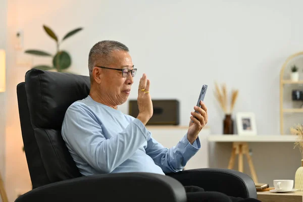 Happy elderly man waving hand, speaking on video call talking to family while resting on armchair at home.