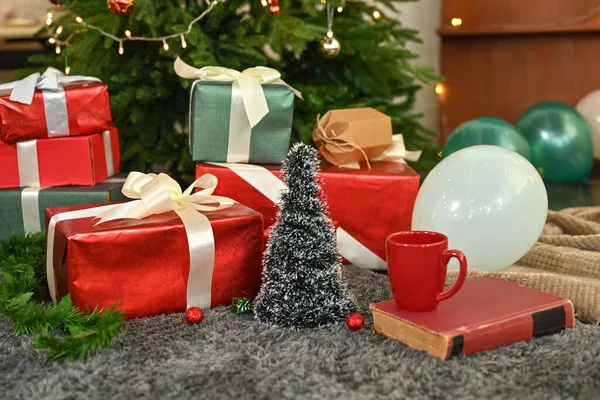 Pile of gift boxes, coffee cup and book on floor near decorated Christmas tree. Winter holiday celebration concept.