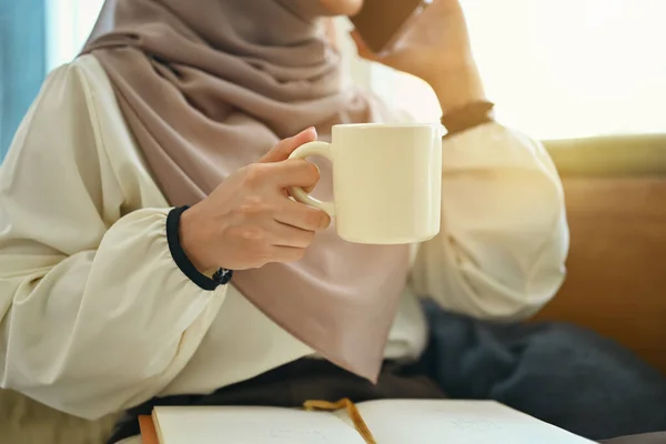 Cropped shot of young muslim woman holding cup of coffee and talking on mobile phone at coffee shop.