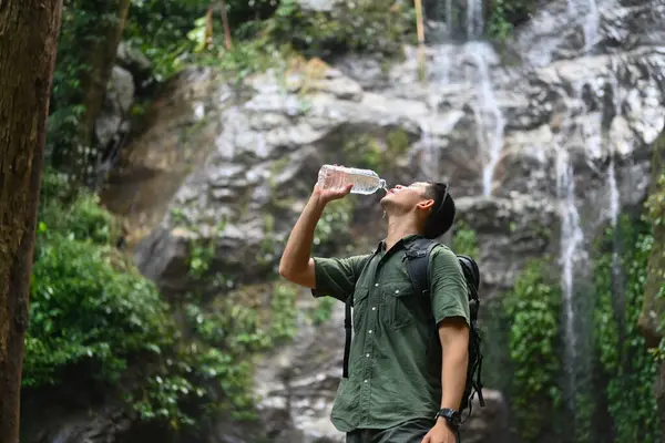 Male hiker with a backpack drinking water from bottle near mountain waterfall.