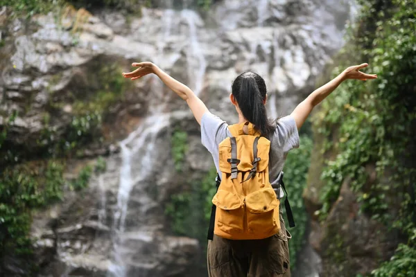 Sırt çantalı kadın gezgin kollarını kaldırarak güzel tropikal şelalenin keyfini çıkarıyor..