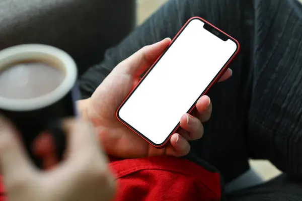 Close up man hands holding smartphone with white screen and drinking coffee on couch.