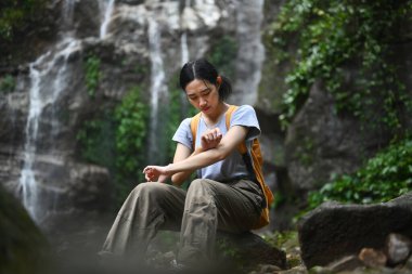 Sırt çantalı kadın yürüyüşçü bir şelalenin önündeki kayanın üzerinde yürüyüş sırasında dinleniyor..