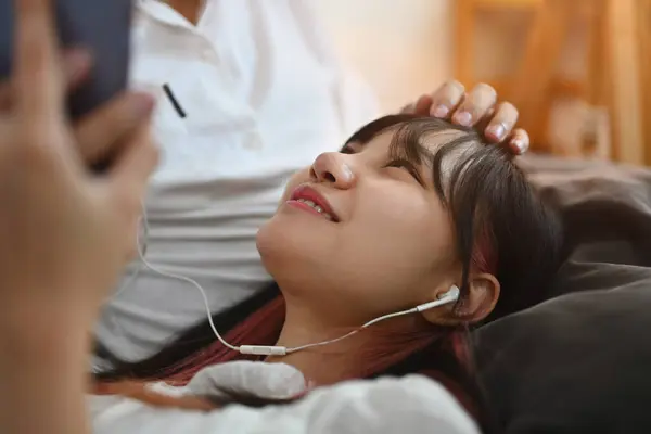 Smiling young woman using mobile phone and listening music new playlist on couch with her boyfriend.