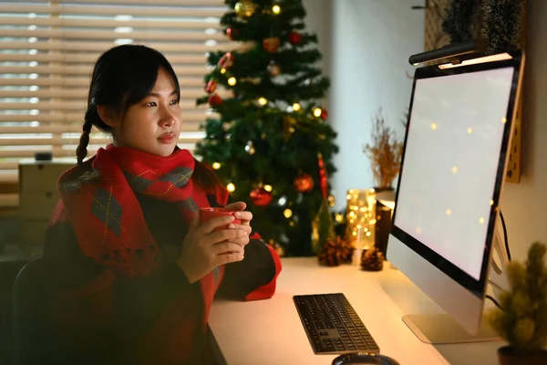 年轻女子拿着一杯热巧克力 看着电脑显示器 背景是圣诞树 — 图库照片