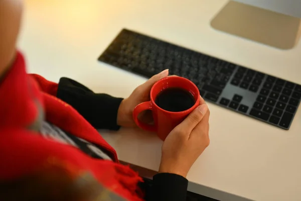 Young woman in warm sweater hands holding cup of hot coffee at white office desk.