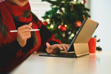 Uyarı giysileri içinde genç bir kadın süslü Noel ağacının yanında oturuyor ve dijital tablet kullanıyor..