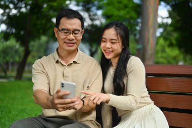 Mutlu genç kadın ve orta yaşlı baba parkta dikilirken cep telefonuyla video izliyorlar..