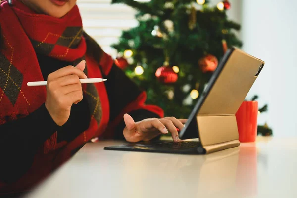 Young woman in warn clothes sitting by decorated Christmas tree and using digital tablet.