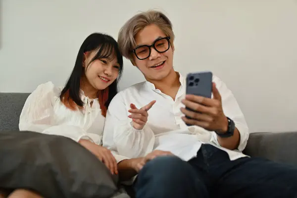 Smiling young couple sharing social media on mobile phone sitting on sofa in living room.