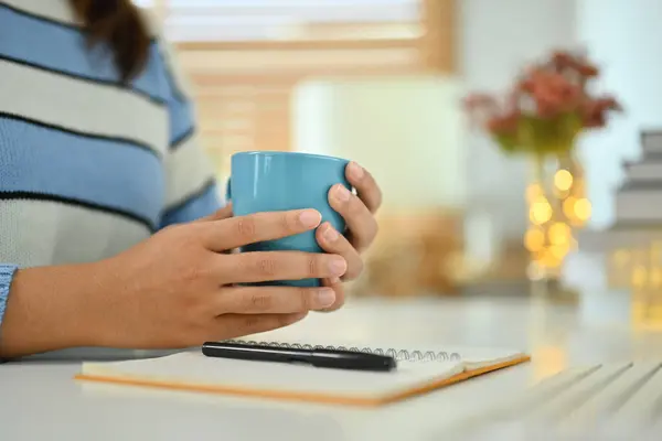 Young woman in warn sweater hands holding cup of herbal tea sitting at workplace.
