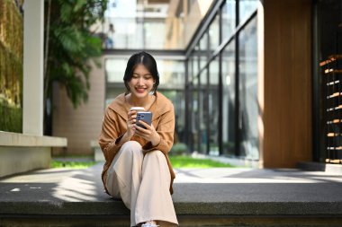 Modern iş binasının önünde otururken cep telefonu kullanan pozitif Asyalı genç bir kadın..