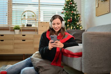 Sosyal medyada cep telefonu sohbeti yapan genç ve güzel bir kadın Noel dekorasyonunda oturma odasında oturuyor..