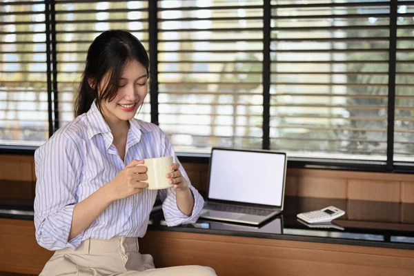 Positive Asian businesswoman taking a break from work and drinking coffee at workplace.