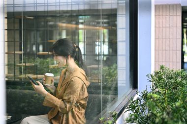 Gülümseyen kadın çalışan kahve içiyor ve modern ofis salonunda cep telefonu kullanıyor..