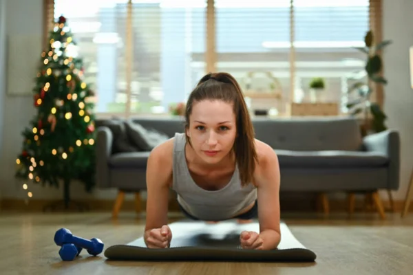 Athletic woman doing plank exercise on mat in living room at Christmas time. Healthy lifestyle concept.