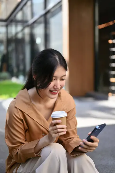 Young businesswoman with take away coffee cup in hand using mobile phone near office building.