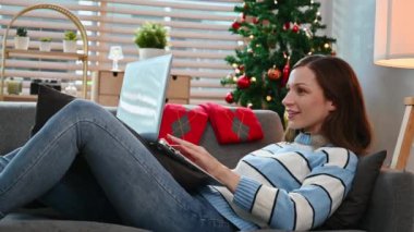 Sıcak kazak giyen, dizüstü bilgisayar kullanan, kış tatillerinde Noel ağacının yanındaki kanepede dinlenen, tatmin olmuş beyaz bir kadın..