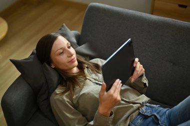 Oturma odasındaki kanepede uzanmış dijital tablette video izleyen rahat genç bir kadın..