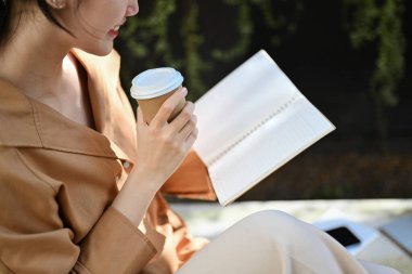 Barışçıl genç kadın kağıt bardaktan kahve içiyor ve açık havada kitap okuyor..