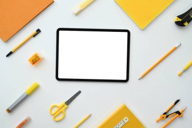 Beyaz arkaplandaki kırtasiyeyle çevrili boş ekranlı dijital tablet. Düz yatış, üst görünüm.
