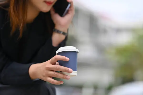 Female worker with paper cup of coffee in hand talking on mobile phone outside office.