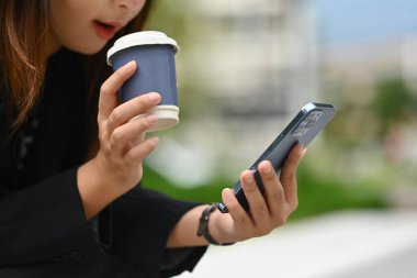 Güneşli bir günde ofis binasının önünde cep telefonuyla kahvesini götüren genç bir kadın..