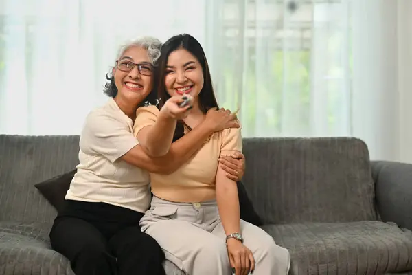 Cheerful retired woman relaxing on sofa with grown up daughter and watching tv on weekend.