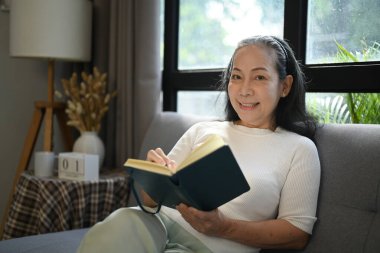 50 'li yaşlardaki gülümseyen kadın günlük kıyafetlerini giymiş evde dinlenirken günlüğünü yazıyor..