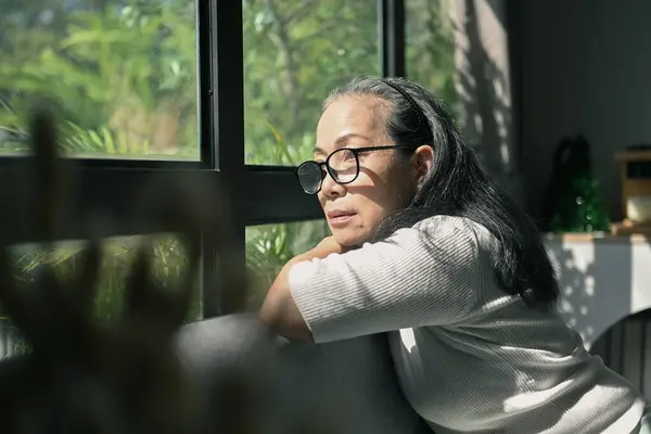 Pensive senior woman sitting on couch and looking in distance out of window