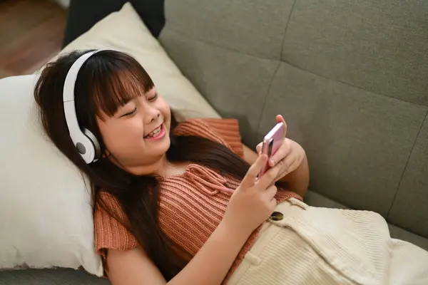 Happy laughing little Asian girl in headphone watching video on mobile phone