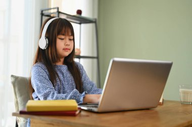 Kulaklıkta konsantre olmuş Asyalı kız evdeki dizüstü bilgisayardan online ders izliyor..